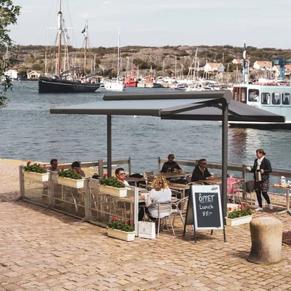 Uteservering vid vattnet med personer som sitter under en terrassmarkis Duo, bakom ligger segelbåtar och en passagerarbåt i Stockholm.