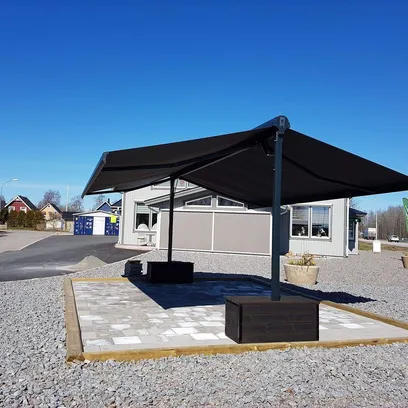 En svart terrassmarkis Duo med fyrkantiga baser placerade på en stenlagd yta utomhus bredvid en grusplan och en grå byggnad under en klar blå himmel i Stockholm.