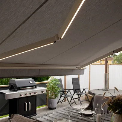 Uteplats med en gasolgrill, sittgrupp med fåtöljer och bord, potted växter och en terrassmarkis med inbyggda LED-lampor i Stockholm.