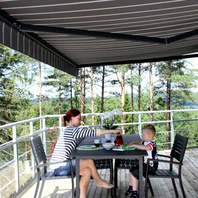 En kvinna och ett litet barn sitter vid ett utebord på en träaltan under en randig terrassmarkis, omgivna av skog i Stockholm.