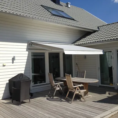 Uteplats med trädäck, ett runt bord med tre trästolar under en vit terrassmarkis T420 på en vit fasad med grått tegeltak och en blå himmel i Stockholm.
