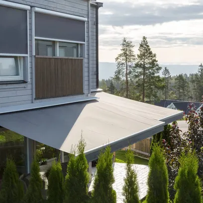 Ett modernt hus med grå träpanel och två utdragna terrassmarkiser T420 över fönster och uteplats, omgiven av gröna träd och buskar i Stockholm.