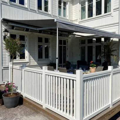 Vit träveranda med vitt staket, terrassmarkis T420 och utemöbler vid ett vitt hus med flera fönster i Stockholm.