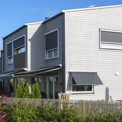 Modern tvåvåningshus med grå träpanel, terrassmarkiser T420, Zipscreen Z460 och en trästaket runt en trädgård i Stockholm.