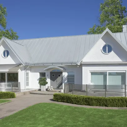 Vit villafasad med två terrassmarkiser T520, två runda fönster under ett plåttak och en grå entrédörr med två cirkulära fönster, omgiven av grön gräsmatta och häckar i Stockholm.