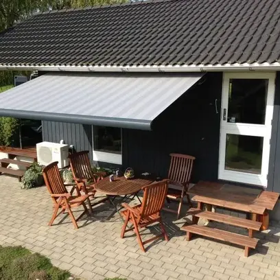 Trädgård med träbord och stolar under en utdragbar terrassmarkis på en stenlagd altan intill ett mörkt hus med vitt dörr- och fönsterkarm i Stockholm.