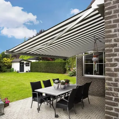 Uteplats med svart-vitrandig terrassmarkis T720, ett bord med sex stolar och blomkrukor på en stenlagd yta intill ett tegelhus med trädgård och lekstuga i bakgrunden i Stockholm.