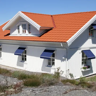 Vit trähus med tegelrött tak och blåa fönstermarkiser F320 på flera fönster, omgiven av klippor och gräsmatta under klarblå himmel i Stockholm.