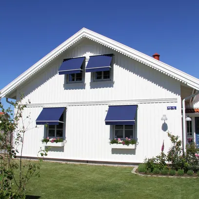 Vit trästuga med blå fönstermarkiser F320 över fönster och välskött gräsmatta under klarblå himmel i Stockholm.