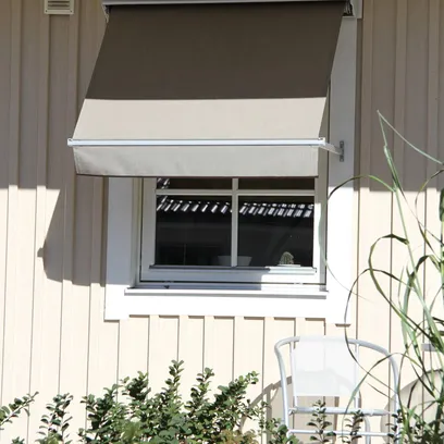 Ett vitt hus med ett fönster täckt av ett brunt och beige fönstermarkis F320, med gröna buskar och en vit stol framför i Stockholm.