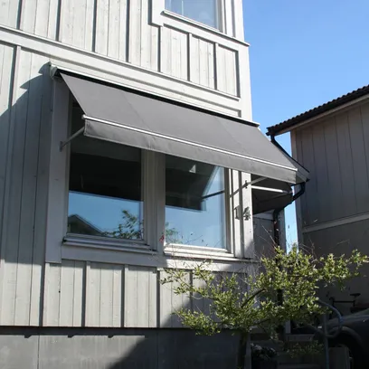 Ett vitt träfasadhus med en grå fönstermarkis F320 över ett fönster och ett litet träd i förgrunden under en klar blå himmel i Stockholm.