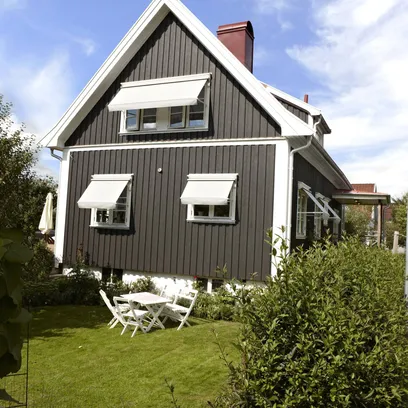 Ett vitt träfasadhus med en grå fönstermarkis F320 över ett fönster och ett litet träd i förgrunden under en klar blå himmel i Stockholm.