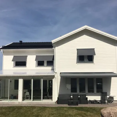 Vit tvåvåningsvilla med grå fönstermarkiser F320 och en terrassmarkis T220, glasat uterum och utemöbler på gräsmatta under blå himmel i Stockholm.