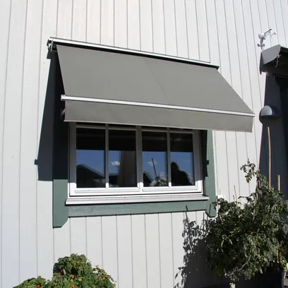 Grå fönstermarkis F320 över ett fönster på vit träpanel med två stora svarta krukor med tomatplantor på vardera sida i Stockholm.