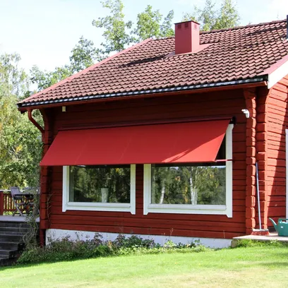 Rött trähus med en röd fönstermarkis F420 över två stora fönster, omgiven av grön gräsmatta och träd i Stockholm.