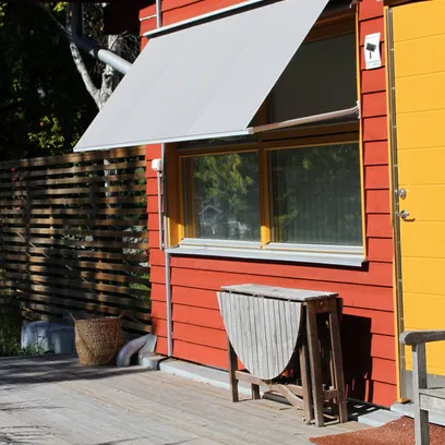 Röd träfasad med ett gult dörr- och fönsterparti, grå fönstermarkis F420 över fönstret, träbord och en rottingkorg på en trätrall i Stockholm.