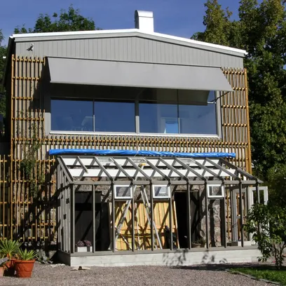 Ett pågående byggprojekt av ett växthus med öppen ramställning framför ett tvåvåningshus med stora fönster och grå fönstermarkis F420 i Stockholm.