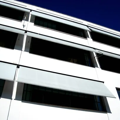 Vitt modernt byggnadsfasad med flera fönster och utsträckta ljusgrå markiser F520 under en klar blå himmel i Stockholm.