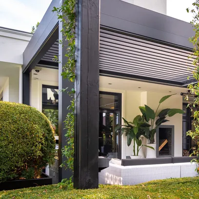 Modernt uterum med pergola Arezzo, vit soffgrupp, stora gröna växter och svart pelare, omgiven av välskötta buskar i Stockholm.