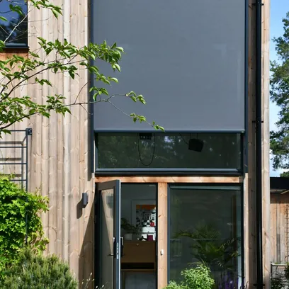 Modern trähus med stora fönster, en av dem har en nedrullad mörkgrå zipscreen Z460 och en dörr står öppen mot en inomhusmiljö i Stockholm.