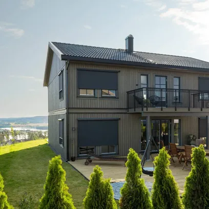 Modernt tvåvåningshus med träfasad, zipscreens, balkong och utomhusmöbler, omgiven av grönska och med utsikt över ett landskap i Stockholm.