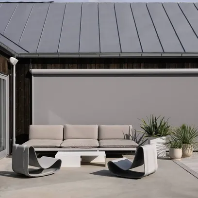 Modern uteplats med beige soffa, två grå loungestolar, vitt soffbord och gröna växter i krukor framför ett hus med metalltak och neddragen markis i Stockholm.