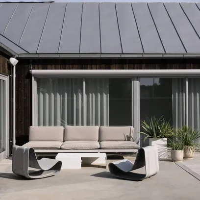 Uteplats med beige soffa, två moderna svarta och vita stolar, vitt soffbord och krukväxter framför ett hus med mörkt träfasad, uppdragen markis och stora glasdörrar i Stockholm.