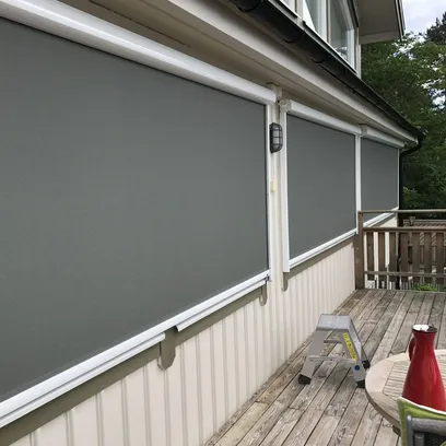 Ett terrassdäck med tre nedrullade grå zipscreens Z560 monterade på husets vita fasad, trästegstege och ett bord med röd kanna i Stockholm.