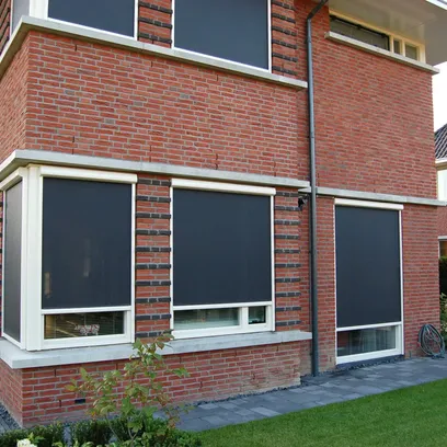 Hörn av ett rött tegelhus med flera fönster täckta av nedrullade mörka zipscreens Z560 och en trädgård med grön gräsmatta och stensatt gångväg i Stockholm.