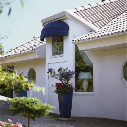 Vit ytterdörr med blå korgmarkis K200 och två höga blå blomkrukor vid ingång till hus med tak i kakel i Stockholm.
