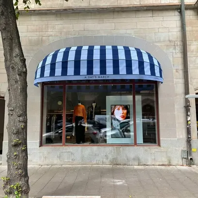 Skyltfönster med randig blå och vit korgmarkis K200 och en skylt med texten A DAY'S MARCH ovanför, visar kläder och ett porträttfoto av en kvinna i Stockholm.