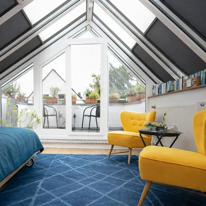 Ljust rum med snedtak i glas, två gula fåtöljer, blå matta och en säng med blått täcke vid en glasdörr till balkong med planteringar och halvt uppdragna takzippar TZ330 i Stockholm.