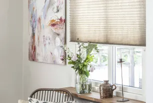 Ett vardagsrum med en halvt nedfälld beige plisségardin, rottingfåtölj med mönstrad kudde, en rund bricka med ljusstakar och böcker, abstrakt konst på väggen, och en fönsterbräda med vas med blommor och keramik i Stockholm.