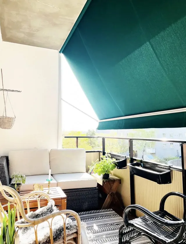 Solig balkong med vita dynor på en hörnsoffa, gröna växter, svart och vit matta samt grön balkongmarkis B260 i Stockholm.