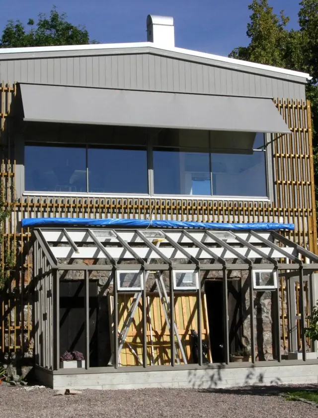 Ett pågående byggprojekt av ett växthus med öppen ramställning framför ett tvåvåningshus med stora fönster och grå fönstermarkis F420 i Stockholm.