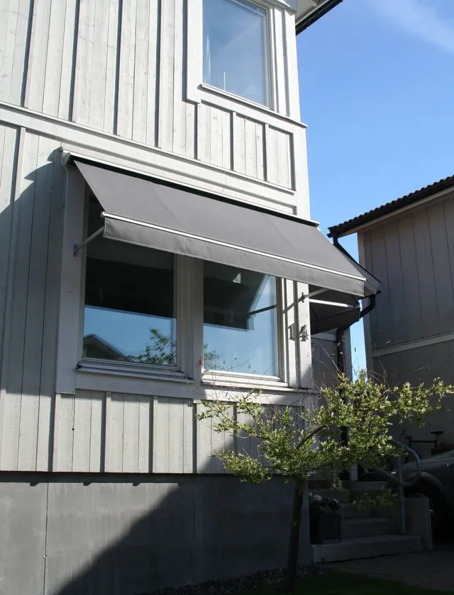 Grå fönstermarkis F320 ovanför två fönster på en vit träfasad med en liten buske framför huset i Stockholm.