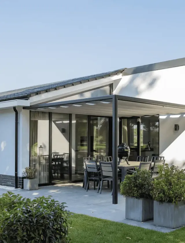 Modern vit villa med skjutdörrar i glas, utemöbler och pergola Sundaze på en stenlagd uteplats med grönt gräs och buskar i Stockholm.