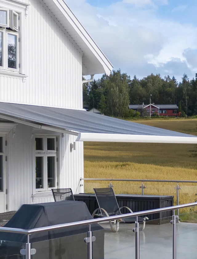 Vit trävilla med utsträckt grå terrassmarkis över en uteplats med svart grill och två svarta stolar, med ett fält och träd i bakgrunden i Stockholm.