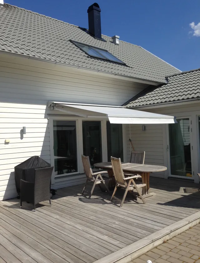 Uteplats med trädäck, ett runt bord med tre trästolar under en vit terrassmarkis T420 på en vit fasad med grått tegeltak och en blå himmel i Stockholm.