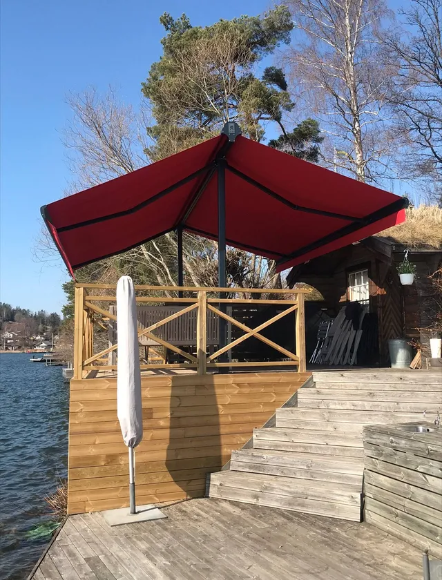 En träaltan vid vattnet med en röd terrassmarkis duo, trappor och ett trähus i bakgrunden under klarblå himmel i Stockholm.