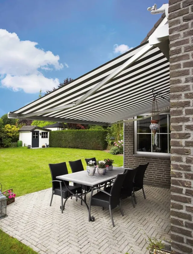 Uteplats med en svartvit randig terrassmarkis, svart bord och sex svarta stolar på en tegelplattad yta vid ett tegelhus i Stockholm.