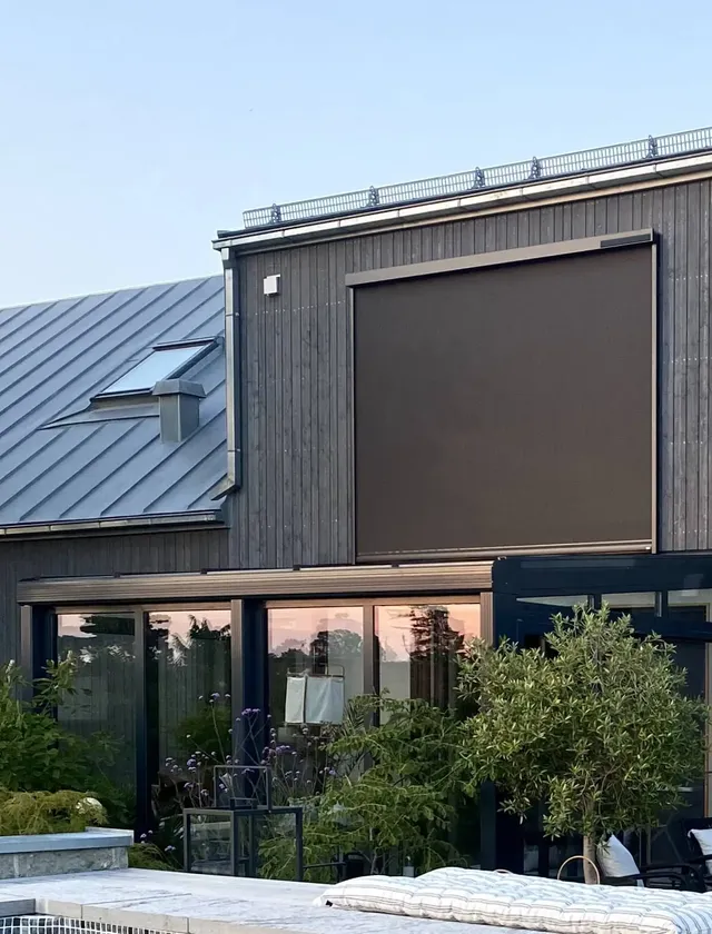 Modernt hus med mörk träfasad, stora fönster och en nedrullad mörk zipscreen Z460 4K på övervåningen, med en pool och växter i förgrunden i Stockholm.
