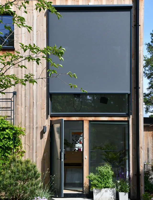 Modern trähus med stora fönster, en av dem har en nedrullad mörkgrå zipscreen Z460 och en dörr står öppen mot en inomhusmiljö i Stockholm.