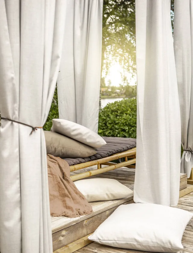En lugn uteplats med en dagbädd med kuddar och gardiner i mjuka beige toner, med grönska och solnedgång i bakgrunden i Stockholm.