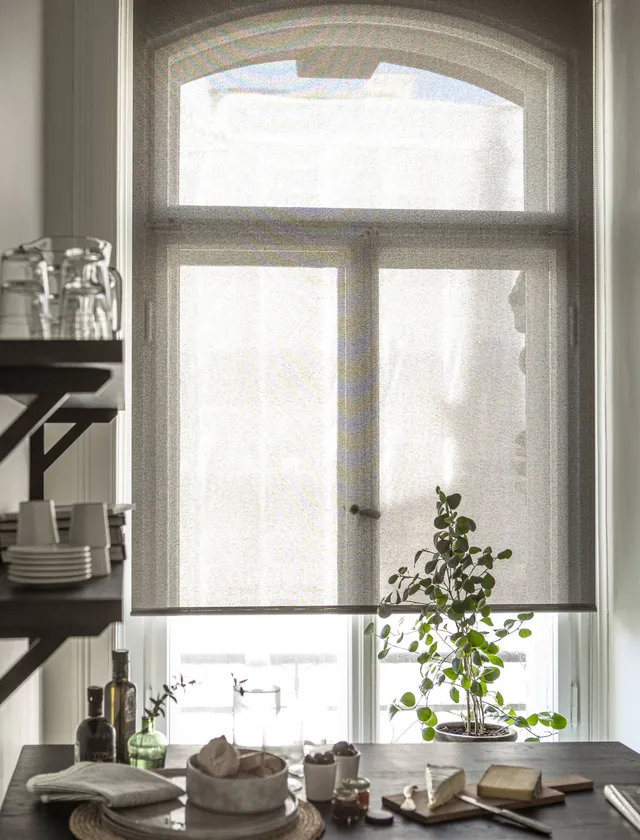Kök med ett fönster täckt av en ljusbeige rullgardin, grönt krukväxt vid fönstret, och ett bord med olika köksartiklar och mat i Stockholm.