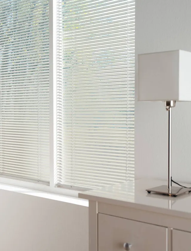 Vita persienner täcker ett fönster bredvid ett bord med en modern bordslampa med vit lampskärm i Stockholm.