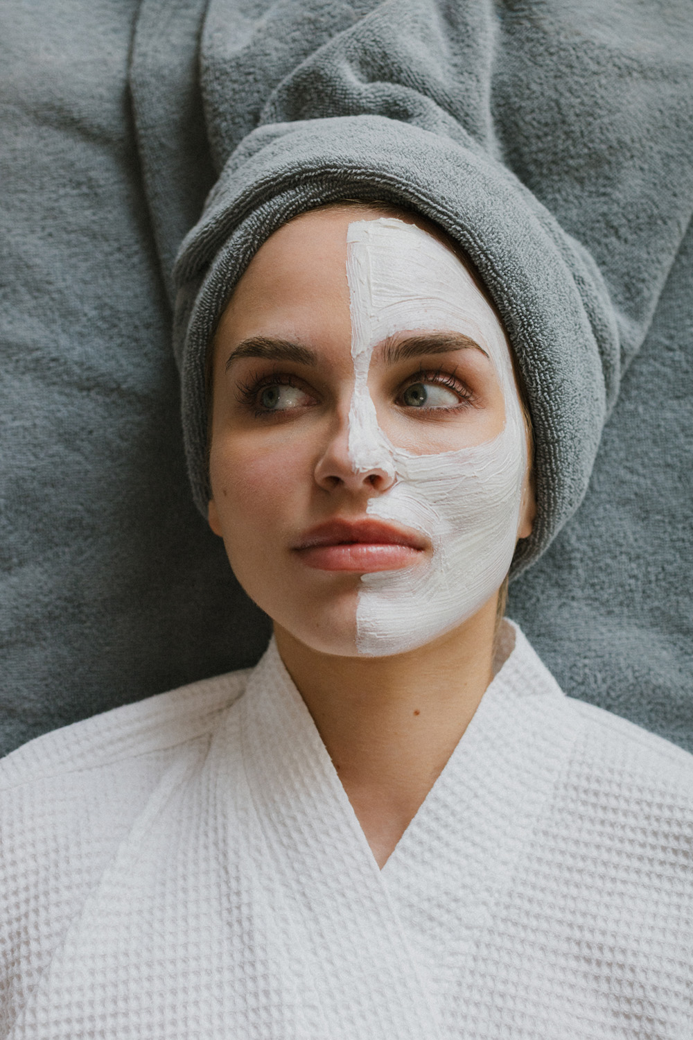 Frau mit grauem Handtuch auf dem Kopf und halbem weißen Gesichtsmaske, die auf grauem Handtuch liegt.