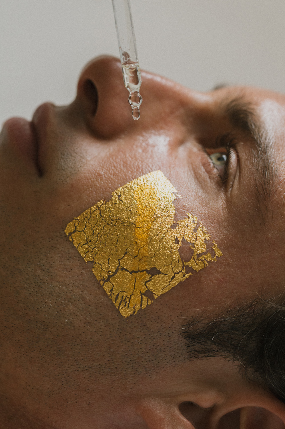 Nahaufnahme eines Mannes mit einem goldenen Blatt auf der Wange und einem Tropfen, der aus einer Pipette fällt.