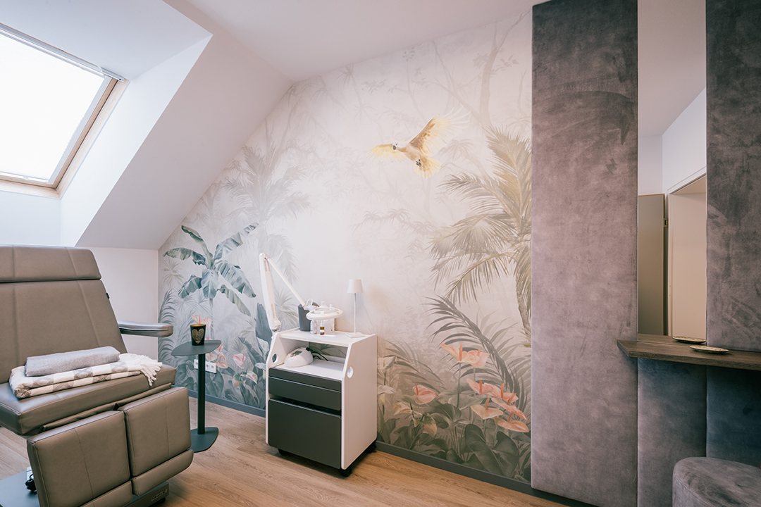 Modernes Behandlungszimmer mit grauem Stuhl, Pflanzenwandmalerei und medizinischem Gerätewagen unter einem Dachfenster.