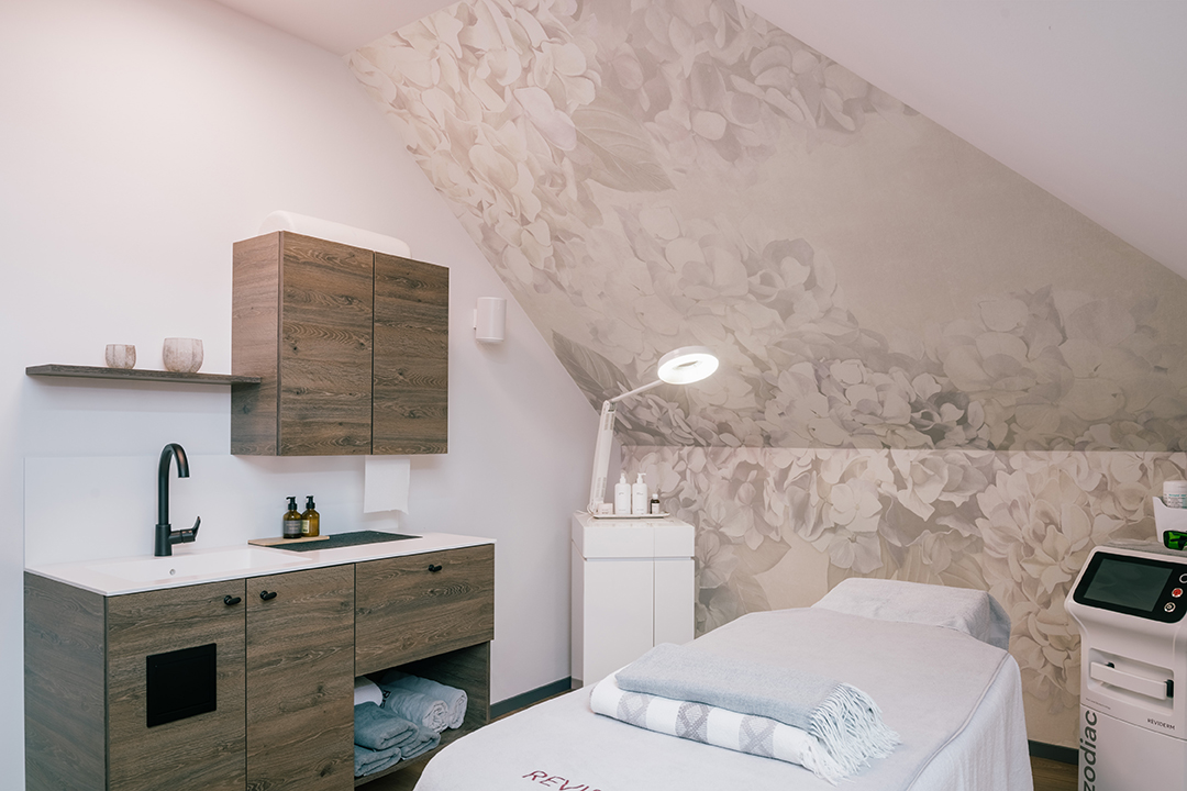 Heller Behandlungsraum mit Massageliege, Holzmöbeln, Waschbecken und großer floral gemusterter Wanddekoration unter einer Dachschräge.